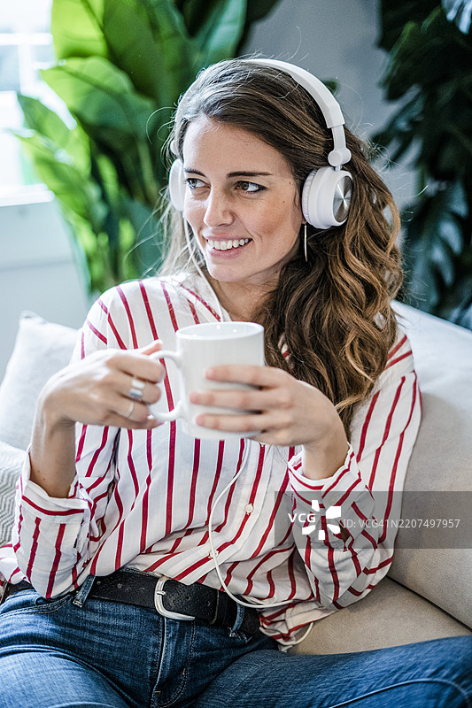 微笑的女性坐在沙发上，手里拿着咖啡杯，戴着耳机。图片素材