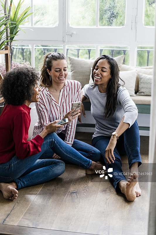 三位快乐的女性坐在家里的地板上，手里拿着手机。图片素材