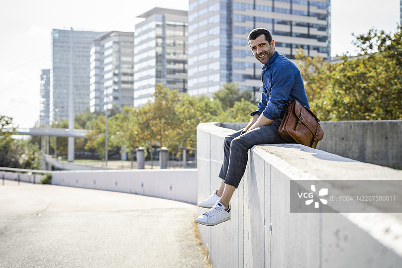 微笑的男子坐在墙上，手里拿着皮革包图片素材