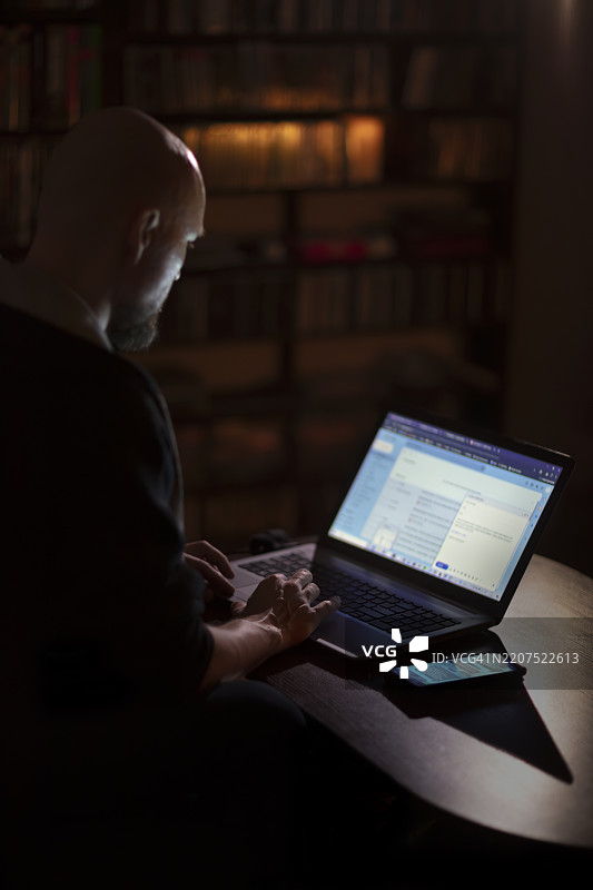 成年男子在家中使用笔记本电脑和手机，夜晚的男性肖像，公寓内。图片素材