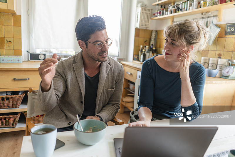 一对情侣坐在厨房的桌子旁，使用笔记本电脑交谈。图片素材