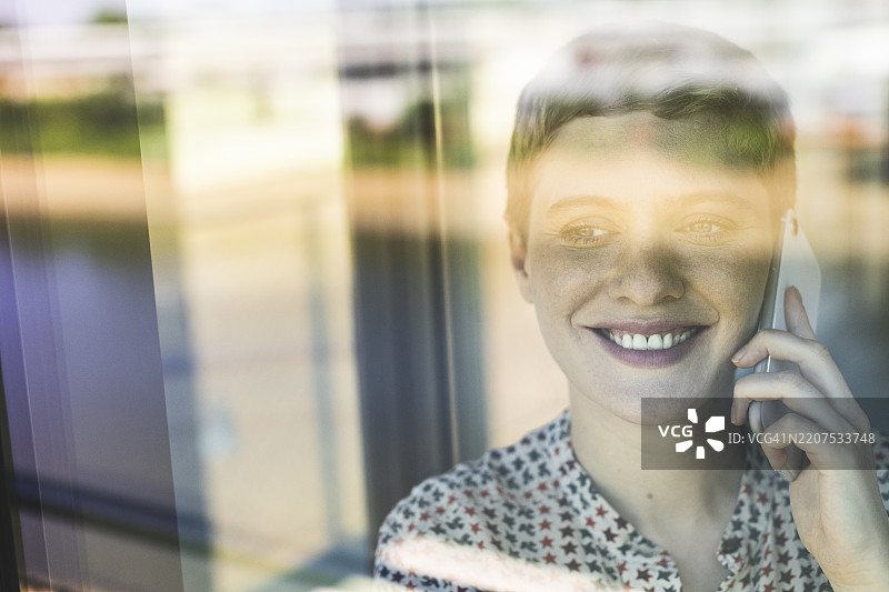 透过窗户看到的微笑女商人在办公室里用智能手机交谈的特写镜头图片素材