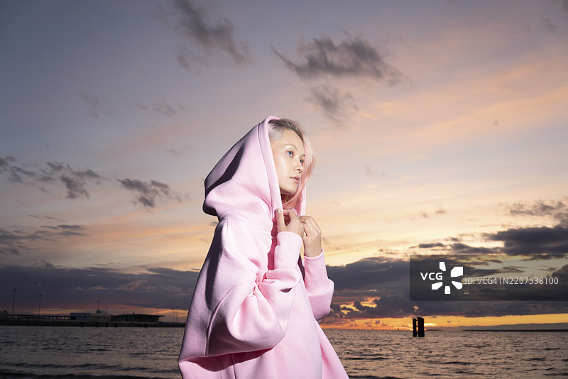 年轻女性穿着粉色连帽衫，站在日落时分的海滩上图片素材
