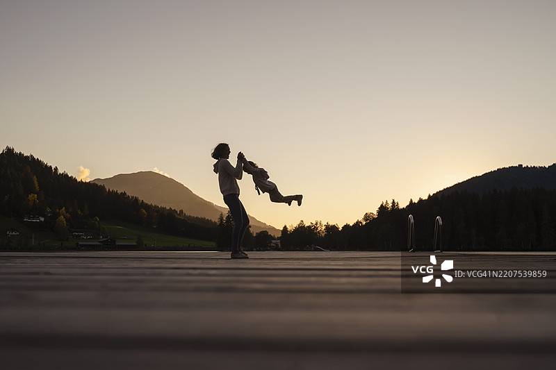 黄昏时分，母亲和小女儿在湖岸码头的剪影中玩耍图片素材