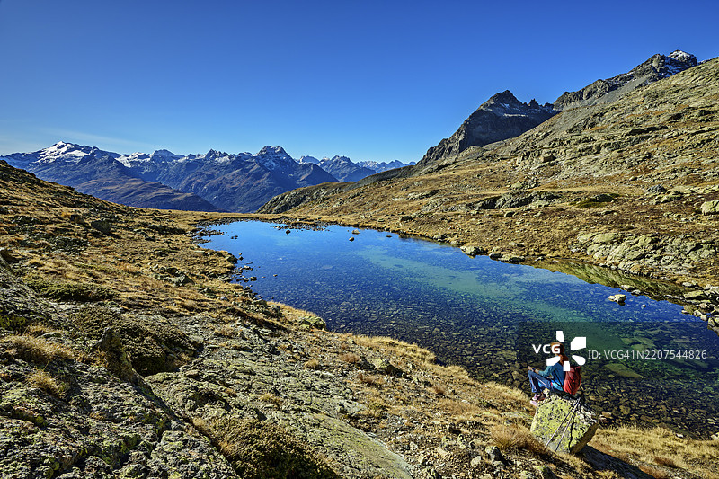 瑞士伯尼纳山脉下，女人坐在湖边的高角度视图，蓝天晴朗。图片素材