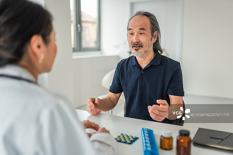 年长的亚洲男性在明亮的办公室里咨询女性医生，桌上有药物。图片素材
