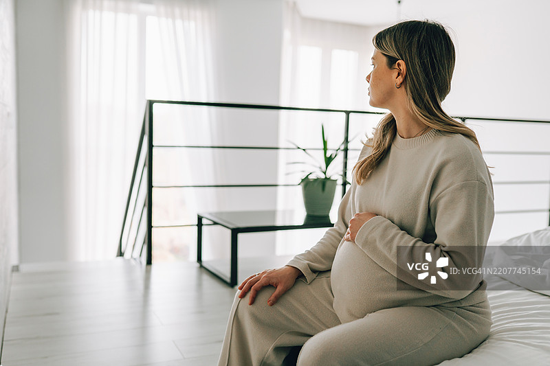 孤独悲伤的孕妇坐在床上，抱着肚子。图片素材