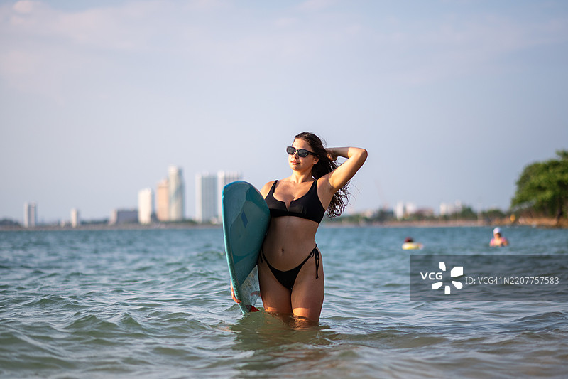 美艳的运动女性在海滩的海岸线上，手里拿着一块蓝色冲浪板，穿着比基尼，场景暗示着温暖的热带或沿海地点，似乎是傍晚或黄昏时分。图片素材