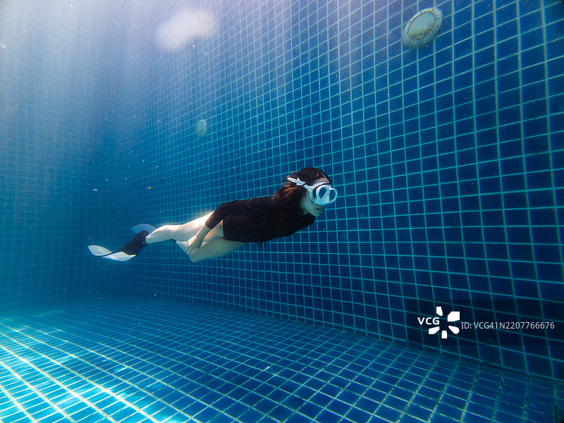 女性潜水者穿着脚蹼和面罩在泳池中游泳，阳光透过水面洒下。图片素材