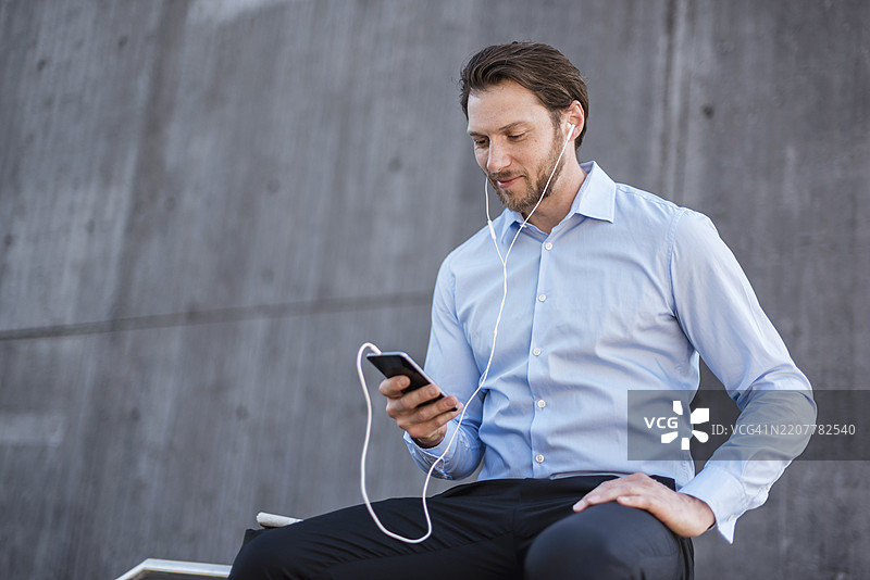 戴着耳机的商人坐在栏杆上看着智能手机图片素材