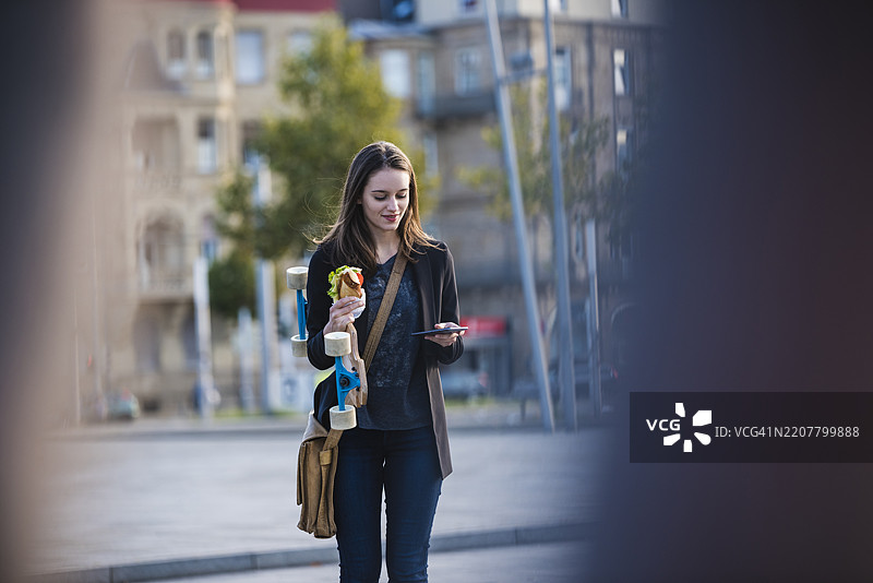 年轻女性在城市里骑着长板，手里拿着零食，查看手机。图片素材