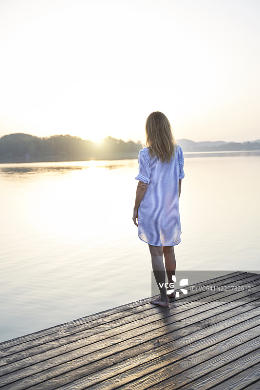 成熟的女性在日出时站在湖边的码头上，背影。图片素材
