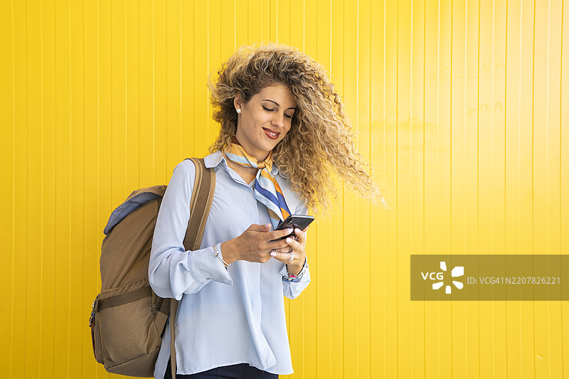 微笑的年轻女性背着背包，站在黄色背景前看着手机。图片素材