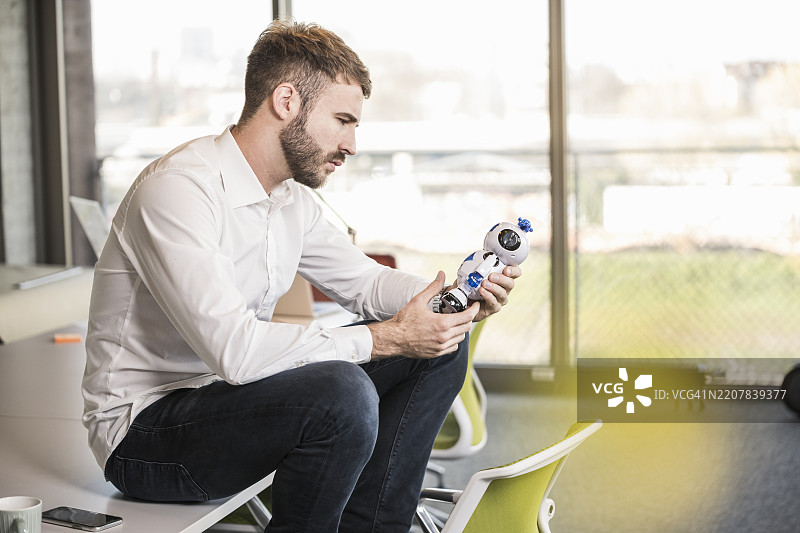 年轻商人在办公室的会议桌上坐着，手里拿着一个机器人。图片素材