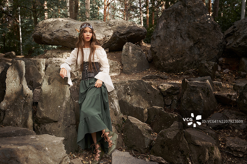 身穿精灵服装的女子正坐在森林中的大石头上。图片素材