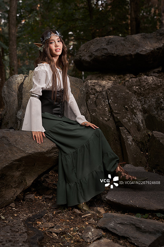 身着精灵服饰的女子正坐在森林中的大石头上。图片素材