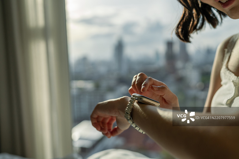 女人坐在床上伸展身体，望向城市天际线，轻触智能手表查看通知。图片素材