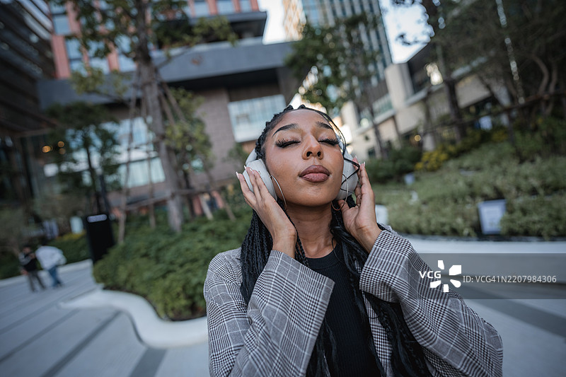 一位黑人女商人在高楼大厦和繁忙城市中，沉浸在无线耳机中的音乐里。图片素材