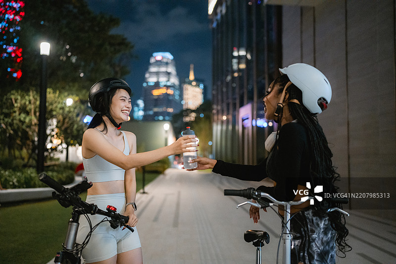 快乐的女性在夜间骑行后将水瓶递给她的朋友们图片素材