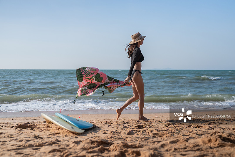 美艳的运动女性在海滩的海岸线上，手里拿着一块蓝色冲浪板。她穿着比基尼，环境暗示着温暖的热带或沿海地点，似乎是傍晚或黄昏时分。图片素材