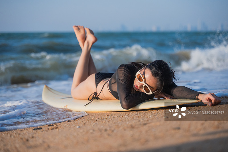 迷人的运动女性在海滩的海岸线上，手里拿着一块蓝色冲浪板。她穿着比基尼，环境暗示着温暖的热带或沿海地点，似乎是傍晚或黄昏时分。图片素材