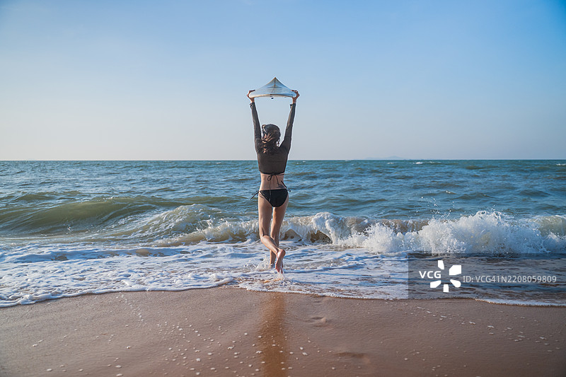 美艳的运动女性在海滩的海岸线上，手里拿着一块蓝色冲浪板。她穿着比基尼，环境暗示着温暖的热带或沿海地点，似乎是傍晚或黄昏时分。图片素材