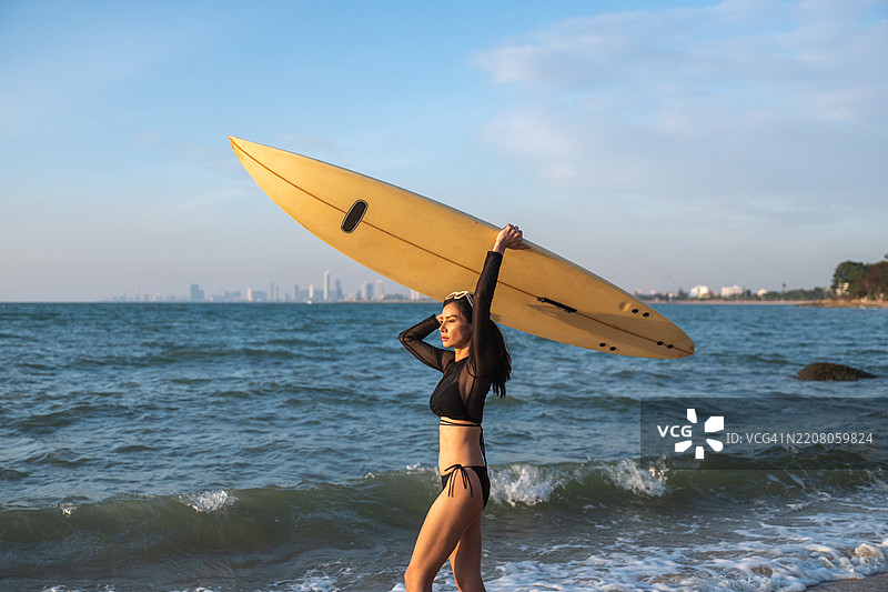 迷人的运动女性在海滩的海岸线上，手里拿着一块蓝色冲浪板，穿着比基尼，场景暗示着温暖的热带或沿海地点，似乎是在傍晚或黄昏时分。图片素材