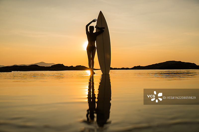 迷人的运动女性在海滩的海岸线上，手里拿着一块蓝色冲浪板，穿着比基尼，场景暗示着温暖的热带或沿海地点，似乎是傍晚或黄昏时分。图片素材