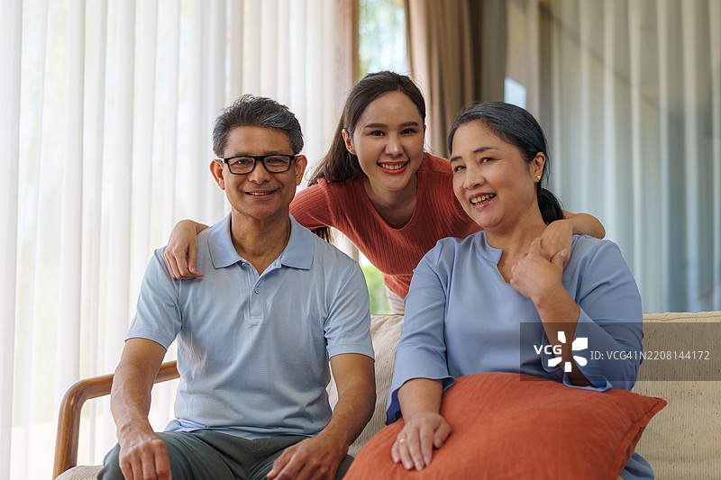 亚洲家庭由父亲、母亲和女儿组成，三人坐在家中的沙发上互相对视并微笑。图片素材
