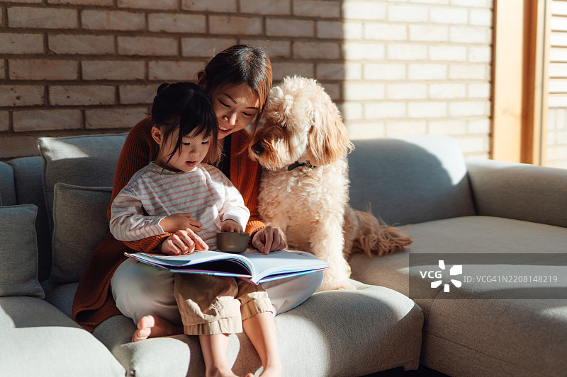 年轻的亚洲母亲在家里的沙发上为可爱的女儿读图画书，身边坐着他们的家庭狗。图片素材