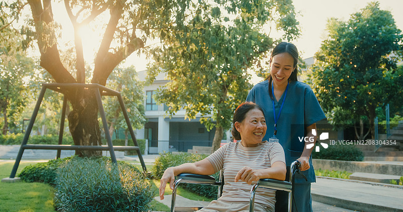 年轻的亚洲护士在公园里推着微笑的老年女性轮椅，传递支持与快乐。户外医疗保健概念。图片素材