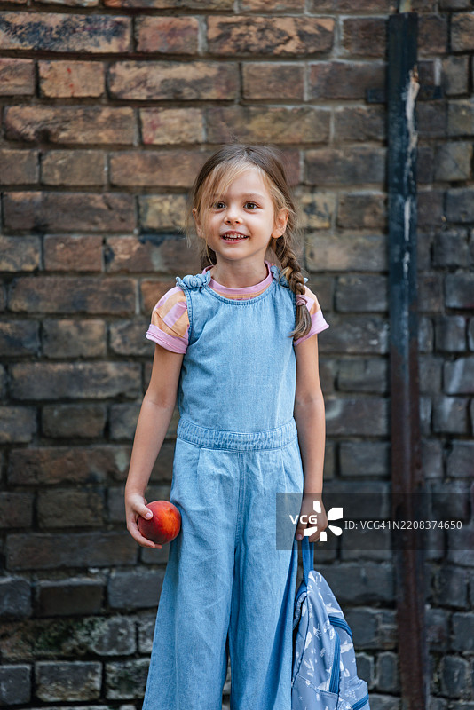 年轻女孩背着书包，手里拿着水果，站在砖墙前图片素材