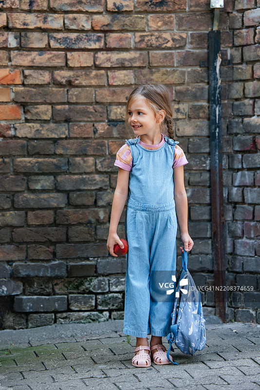 微笑的年轻女孩手持苹果和背包，站在砖墙前图片素材