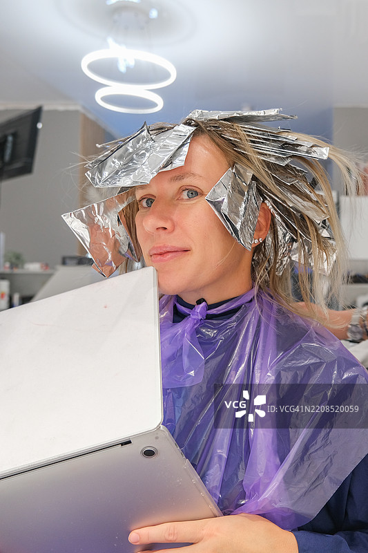 一个女孩在沙龙里染头发的同时，使用电脑远程工作。图片素材