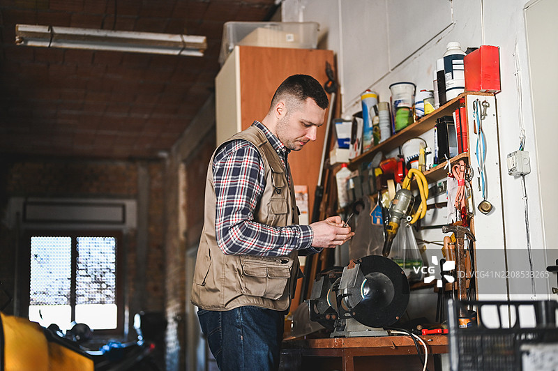 机械师在他的工作坊里使用台式磨床磨锐工具，周围是各种工具和设备。图片素材
