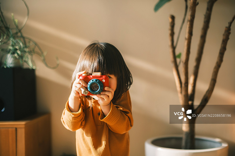 可爱的微笑小亚洲女孩像专业摄影师一样，开心地用木制玩具相机在温暖的阳光下的舒适家中拍照。孩子们的游戏时间。鼓励创造力和想象力。通过游戏学习。图片素材