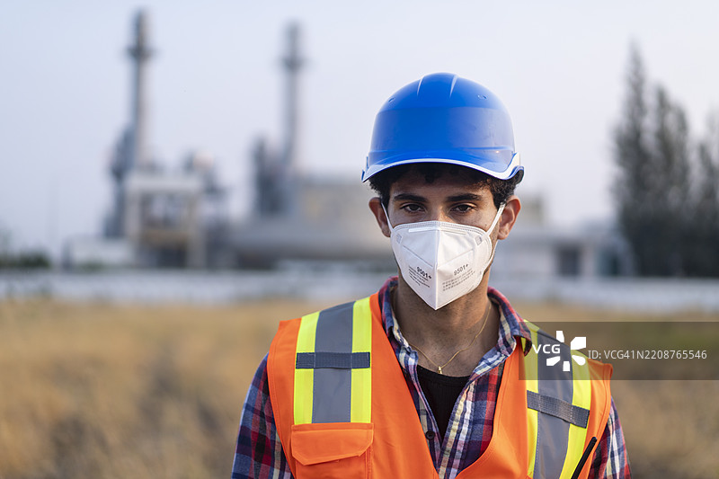 戴着口罩和安全头盔的工程师站在石油炼油厂和石化厂前，背景有污染。图片素材