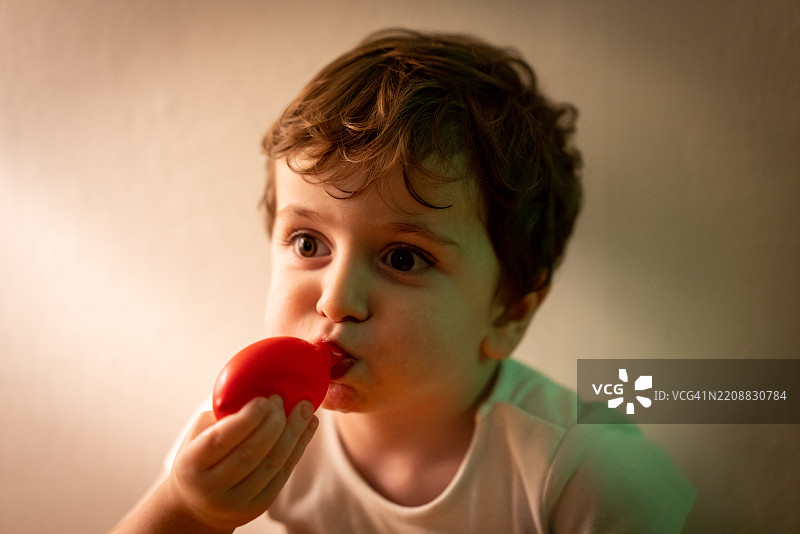 小男孩独自坐在白墙旁，嘴里吹着气球。图片素材