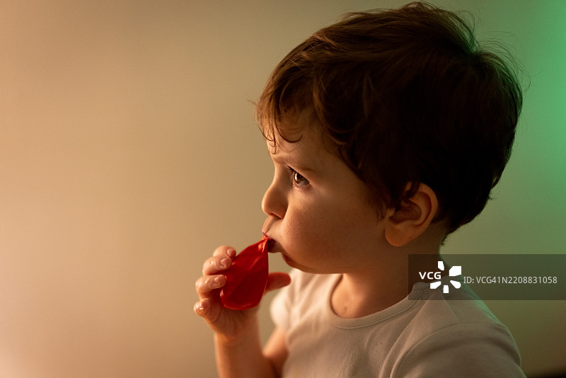 小男孩独自坐在白墙旁，嘴里吹着气球。图片素材