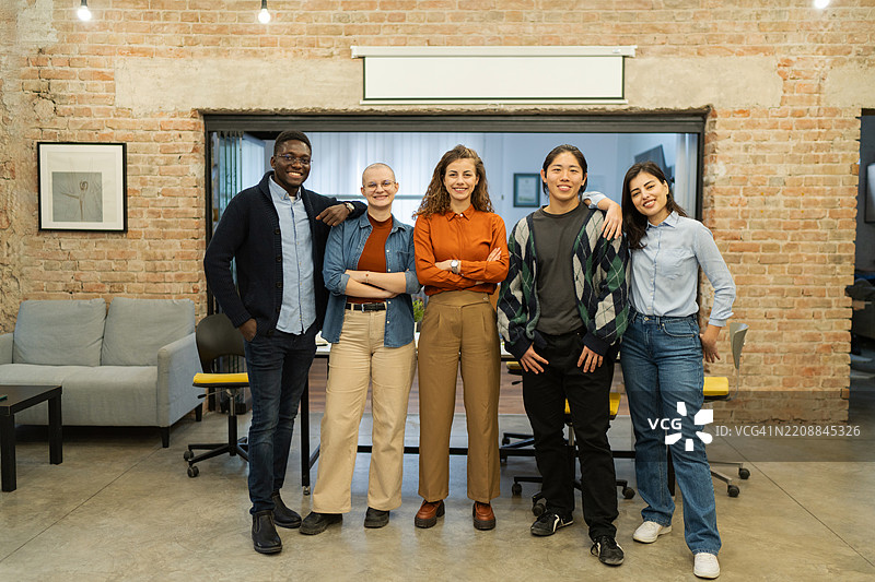 一群五位年轻的商业人士在现代共享办公空间中站在一起，自信地对着镜头微笑。图片素材