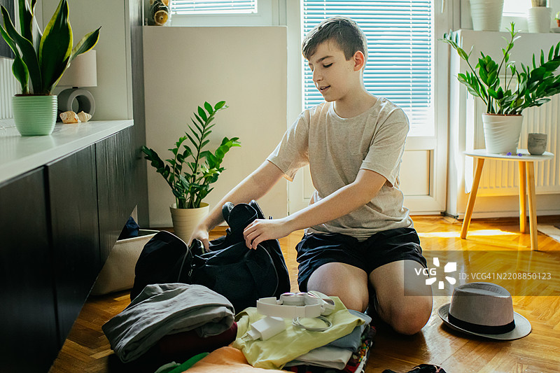 准备即将到来的夏季旅行。快乐的少年在家里的地板上为家庭度假或学校远足打包旅行包。图片素材