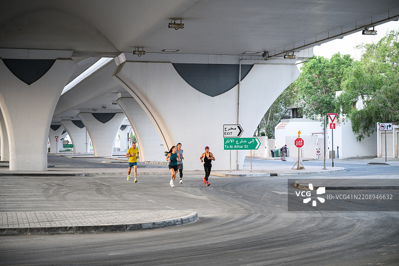 远处广角镜头拍摄的慢跑者在城市桥下奔跑，背景是几何现代建筑和混凝土纹理。图片素材