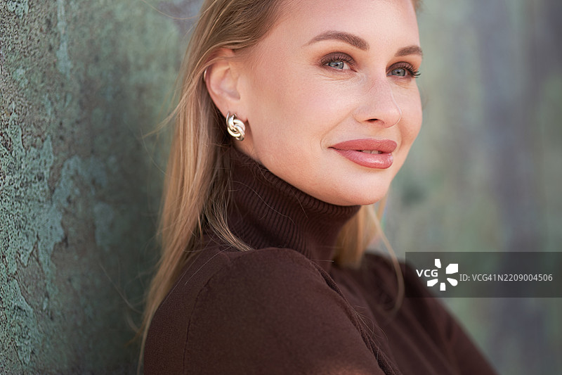 一位金发微笑的女性，穿着棕色高领毛衣，耳朵上挂着耳环。图片素材