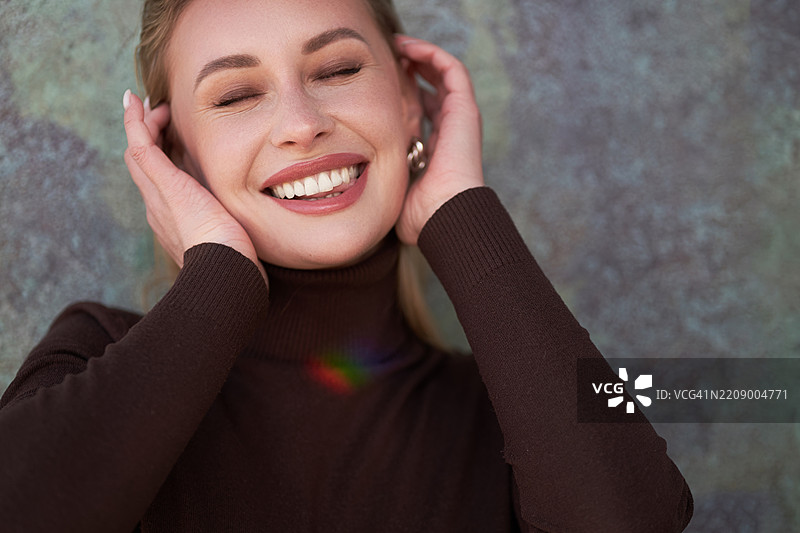 穿着棕色高领毛衣的快乐女性。她双手托着脸，闭着眼睛微笑。图片素材