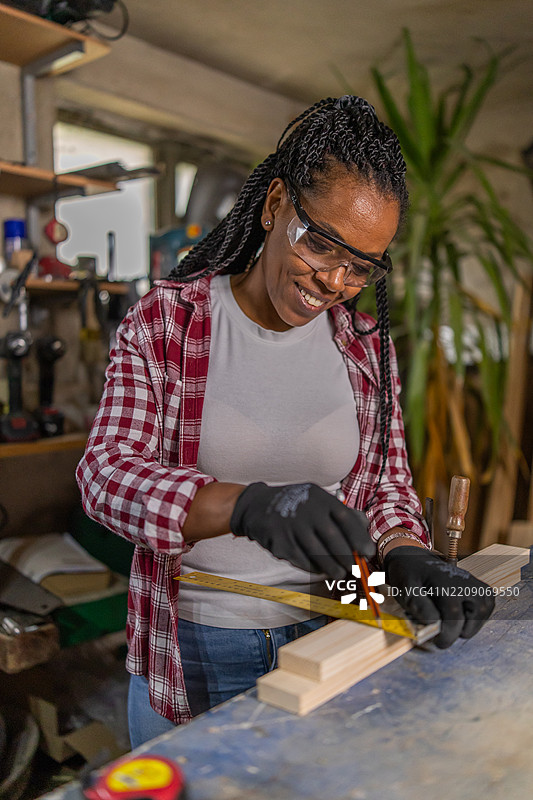 African female with protective equipment measuring plank with measure meter in carpentry work shop.图片素材