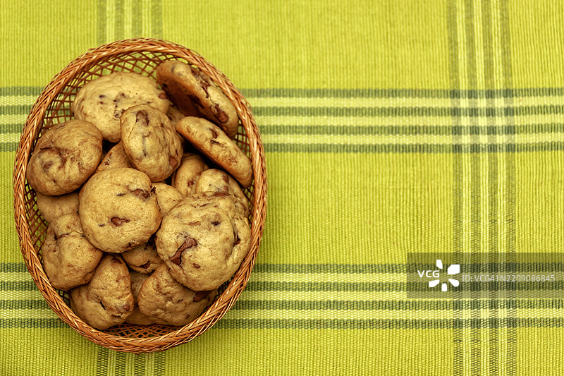 从上方看到的画面。左侧是一个特写，展示了放在绿色格子布上的自制巧克力碎片蜂蜜饼干，放在一个篮子里。留有空白空间。图片素材