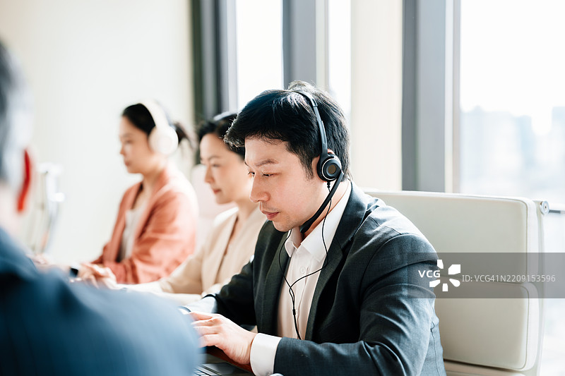 亚洲商人佩戴耳机与坐在会议室的团队进行视频会议。图片素材