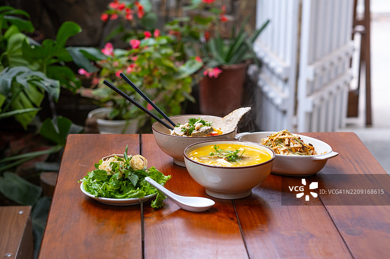 一组越南菜肴摆放在木桌上，背景有绿植和花朵。包括米粉汤、香草、豆腐和配料。图片素材