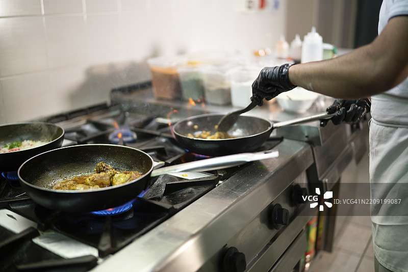 专业厨师戴着黑色手套，熟练地在炉子上的平底锅中混合食材，周围是蓝色火焰，身处繁忙的商业厨房环境中。图片素材