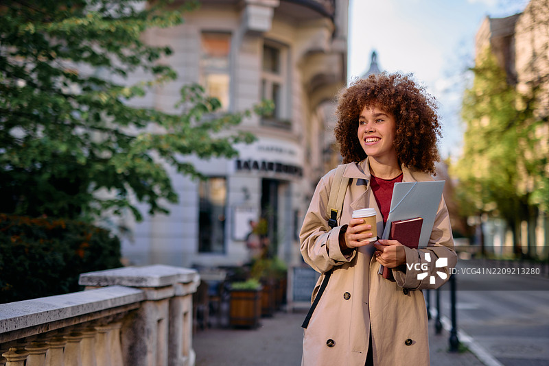 微笑的学生在城市中走着，手里拿着书和咖啡图片素材
